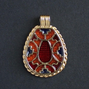 May include: A gold-toned pendant with a teardrop shape, featuring a red and orange enamel design with navy blue accents. The pendant has a rope-like border and a gold bail for a chain.