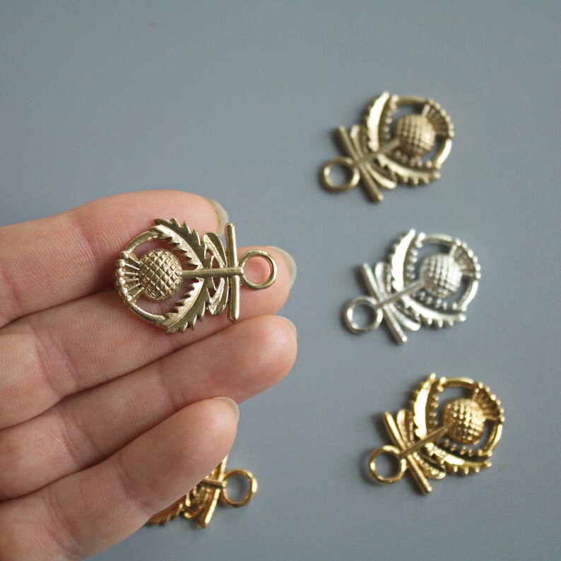 May include: Four gold-toned metal charms featuring a stylized thistle design. The charms have a loop at the top and a toggle clasp on the side.