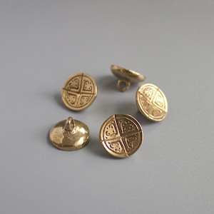 May include: Five antique brass buttons with a cross-like design. Each button has a loop on the back for sewing. The buttons are round and have a slightly domed shape. The buttons are a warm gold color.