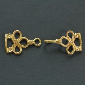 Può includere: Una chiusura dorata con un design intricato ad anello. La chiusura è composta da due parti: una sezione ad anello con un anello e un pezzo a forma di gancio. Il design presenta una serie di anelli interconnessi ed elementi decorativi.