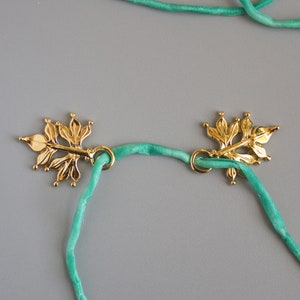 Pu&ograve; includere: Due chiusure dorate a forma di foglia, collegate da un cordoncino verde chiaro. Le chiusure presentano un design floreale dettagliato e sono poste su uno sfondo grigio. Il cordoncino sembra essere fatto di un materiale morbido e setoso.
