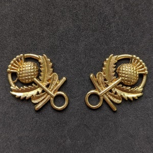 May include: Two gold-toned metal toggle clasps with a thistle design. Each clasp features a stylized thistle with leaves and a ring for attaching to a garment.
