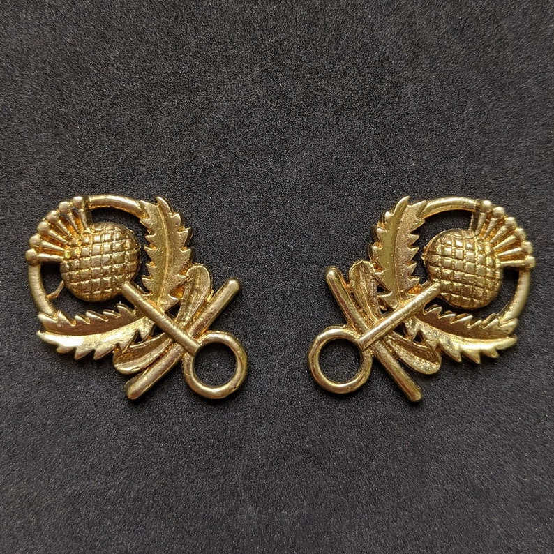 May include: Two gold-toned metal toggle clasps with a thistle design. Each clasp features a stylized thistle with leaves and a ring for attaching to a garment.