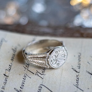 May include: A silver signet ring with a detailed design of a stylized flower and two swords. The ring has a beaded edge and a textured band.