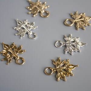 Pu&ograve; includere: Primo piano di diversi ciondoli metallici a forma di foglia in tonalit&agrave; oro e argento. Ogni ciondolo presenta un design floreale dettagliato con un piccolo anello per il fissaggio a gioielli o altri lavori artigianali. I ciondoli sono disposti su una superficie grigia.