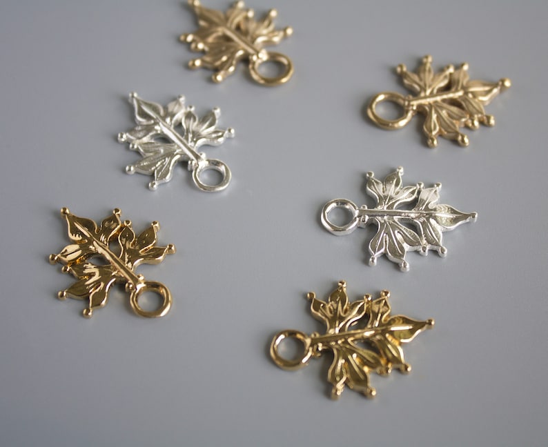 Pu&ograve; includere: Primo piano di diversi ciondoli metallici a forma di foglia in tonalit&agrave; oro e argento. Ogni ciondolo presenta un design floreale dettagliato con un piccolo anello per il fissaggio a gioielli o altri lavori artigianali. I ciondoli sono disposti su una superficie grigia.