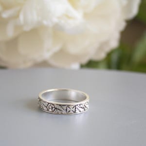 Pu&ograve; includere: Un anello a fascia d'argento con un design floreale e geometrico dettagliato. L'anello &egrave; su una superficie grigia.
