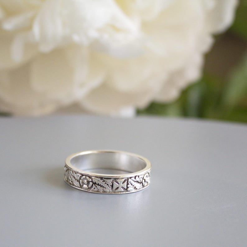 Pu&ograve; includere: Un anello a fascia d'argento con un design floreale e geometrico dettagliato. L'anello &egrave; su una superficie grigia.