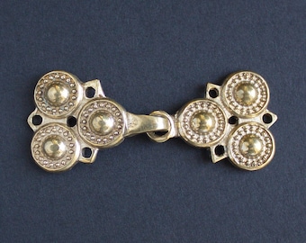 Puede incluir: Un cierre de latón de estilo vintage con un diseño decorativo. Cuenta con múltiples elementos circulares con patrones de puntos en relieve, conectados por una barra central y un anillo. El metal tiene una apariencia cálida y envejecida, lo que sugiere una estética histórica o antigua.