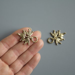 Pu&ograve; includere: Due elementi per gioielli a forma di foglia color ottone, con un anello per il fissaggio. Gli elementi hanno un design botanico dettagliato con piccoli accenti arrotondati. Lo sfondo &egrave; grigio neutro.