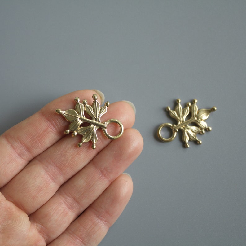 Pu&ograve; includere: Due elementi per gioielli a forma di foglia color ottone, con un anello per il fissaggio. Gli elementi hanno un design botanico dettagliato con piccoli accenti arrotondati. Lo sfondo &egrave; grigio neutro.