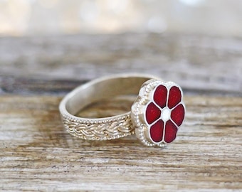 May include: A silver ring with a red flower-shaped design. The flower has six petals and a white center. The band features a braided pattern. The ring is set against a blurred wooden background.