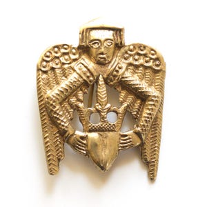 May include: A gold-toned brooch featuring a detailed design of a figure with wings, holding a crown and a heart-shaped shield. The brooch has intricate detailing and a pin closure on the back. The overall design is reminiscent of heraldic imagery.