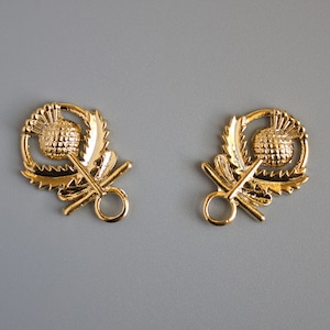 May include: Two gold-toned metal thistle-shaped clasps with a ring on each side. The clasps are detailed with a raised thistle design.