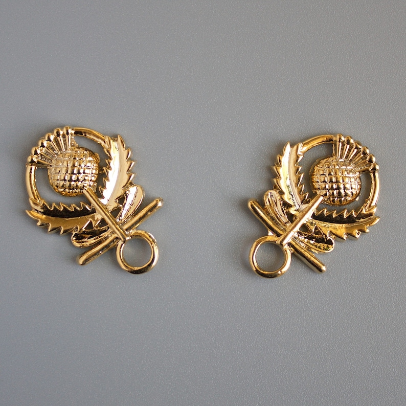 May include: Two gold-toned metal thistle-shaped clasps with a ring on each side. The clasps are detailed with a raised thistle design.