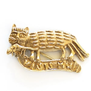 Op de afbeelding: Goudkleurige kattenbroche met een gestileerd ontwerp. De kat houdt een vis vast en staat op een banner met de tekst "VISIS MU". De broche heeft een speld op de achterkant voor bevestiging.