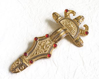 May include: A gold-colored brooch with intricate designs and red gemstones. The brooch is shaped like a sword with a decorative handle.