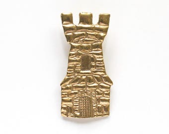 Puede incluir: Broche dorado con forma de torre de castillo. El broche presenta un diseño detallado de una torre de castillo con almenas en la parte superior y una pequeña puerta en la base. La superficie tiene un aspecto texturizado similar a la piedra.