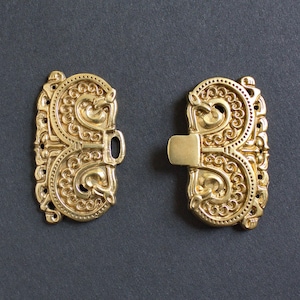 Peut inclure: Deux fermoirs dorés ornés avec des volutes complexes et un mécanisme de verrouillage central. Les fermoirs sont symétriques, avec un design détaillé de courbes et de motifs. Le fond est de couleur gris foncé uni.