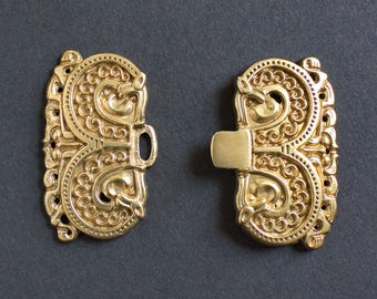 May include: Two ornate gold-colored clasps with intricate scrollwork and a central locking mechanism. The clasps are symmetrical, with a detailed design featuring curves and patterns. The background is a solid, dark gray color.