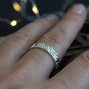 Pu&ograve; includere: Un anello a fascia d'argento con un intricato design floreale. L'anello &egrave; indossato su un dito.