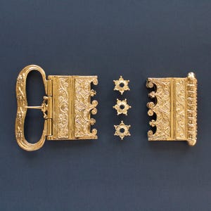 Puede incluir: Cierre dorado ornamentado con intrincados diseños florales y de volutas. El cierre está abierto, revelando sus componentes, incluyendo tres pequeños elementos decorativos en forma de estrella. El fondo es azul oscuro sólido.