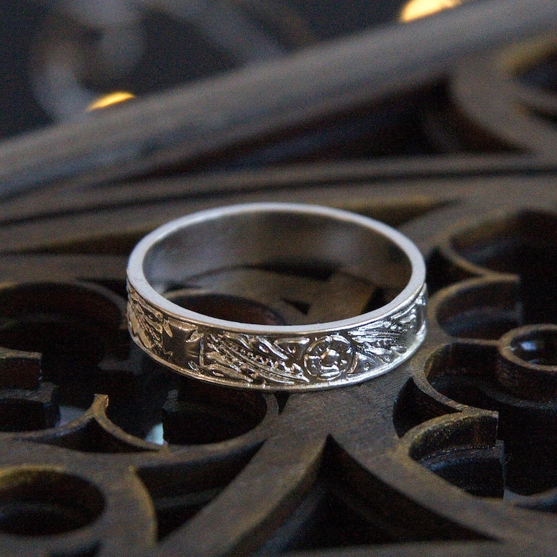 Pu&ograve; includere: Un anello a fascia d'argento con un intricato design floreale. L'anello &egrave; su una superficie di legno scuro.