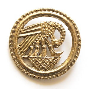 May include: A gold-colored, circular brooch with an intricate design. The brooch features a central motif of a stylized animal or mythical creature within a beaded border. The design is detailed and appears to be vintage or antique.