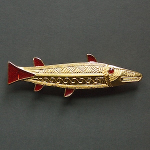 May include: A gold-toned fish-shaped brooch with red enamel accents on the fins and tail. The brooch features intricate engraved patterns and a red gemstone eye, showcasing a detailed design. The fish is approximately 3 inches long.