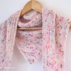 May include: A pale pink, speckled knitted scarf hangs on a wooden clothes hanger. The scarf has a triangular shape and is made of a soft, textured yarn with flecks of colour. A cosy accessory.