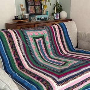 May include: A colorful, square crochet blanket draped over a sofa. The blanket features a geometric pattern with stripes of blue, green, pink, and white. The blanket is made of soft yarn and is perfect for adding a cozy touch to any living space.