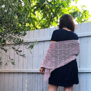 Puede incluir: Un chal de ganchillo rosa con un patrón de encaje y pequeños pompones rosas. El chal está drapeado sobre los hombros de una persona que lleva un vestido negro.