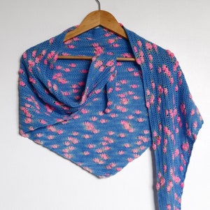 May include: A blue crochet shawl with a pink floral pattern. The shawl is draped over a wooden hanger.