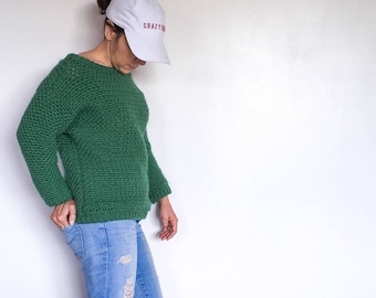 May include: A woman wearing a green knitted sweater, blue jeans with distressed details, and white sneakers with pink stripes. She is wearing a white baseball cap with the text "CRAZY" in pink.