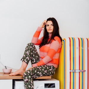 May include: A woman wearing a pink and orange striped sweater and floral print pants sits on a washing machine. A colorful striped refrigerator is to the right. A small pink bowl sits on the countertop.