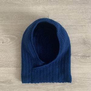 Pode incluir: Um cachecol de crochê azul escuro com capuz. O cachecol é feito com um padrão de ponto simples e tem um capuz largo e aberto.