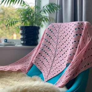 May include: A pink crocheted shawl draped over a teal armchair. The shawl has a delicate openwork pattern and is made with a soft yarn.
