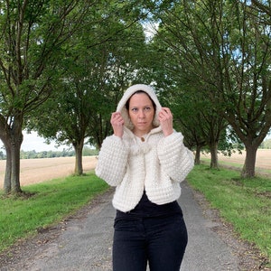 May include: A white crocheted hooded cardigan with a tassel detail. The cardigan is worn by a person standing on a path lined with trees.