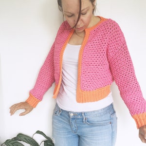 May include: A pink crocheted cardigan with orange trim. The cardigan is cropped and has a V-neck.