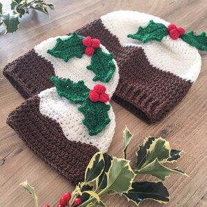 May include: Two crocheted Christmas pudding hats with green holly and red berry embellishments. The hats are brown and white and are sitting on a wooden surface with green holly leaves.