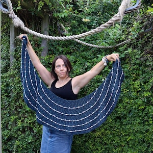 May include: A teal blue crocheted shawl with white and pink beads. The shawl is draped over the shoulders of a person standing in front of a green leafy background.