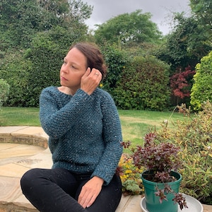 May include: A woman wearing a teal blue sweater with a speckled pattern. She is sitting on a stone patio with green grass and plants in the background.
