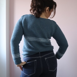 Dora is seen from behind wearing a blue long sleeve relaxed fit crochet sweater made from the Sequel sweater pattern. She has her hands on her hips, showing the stripes around the wrist.