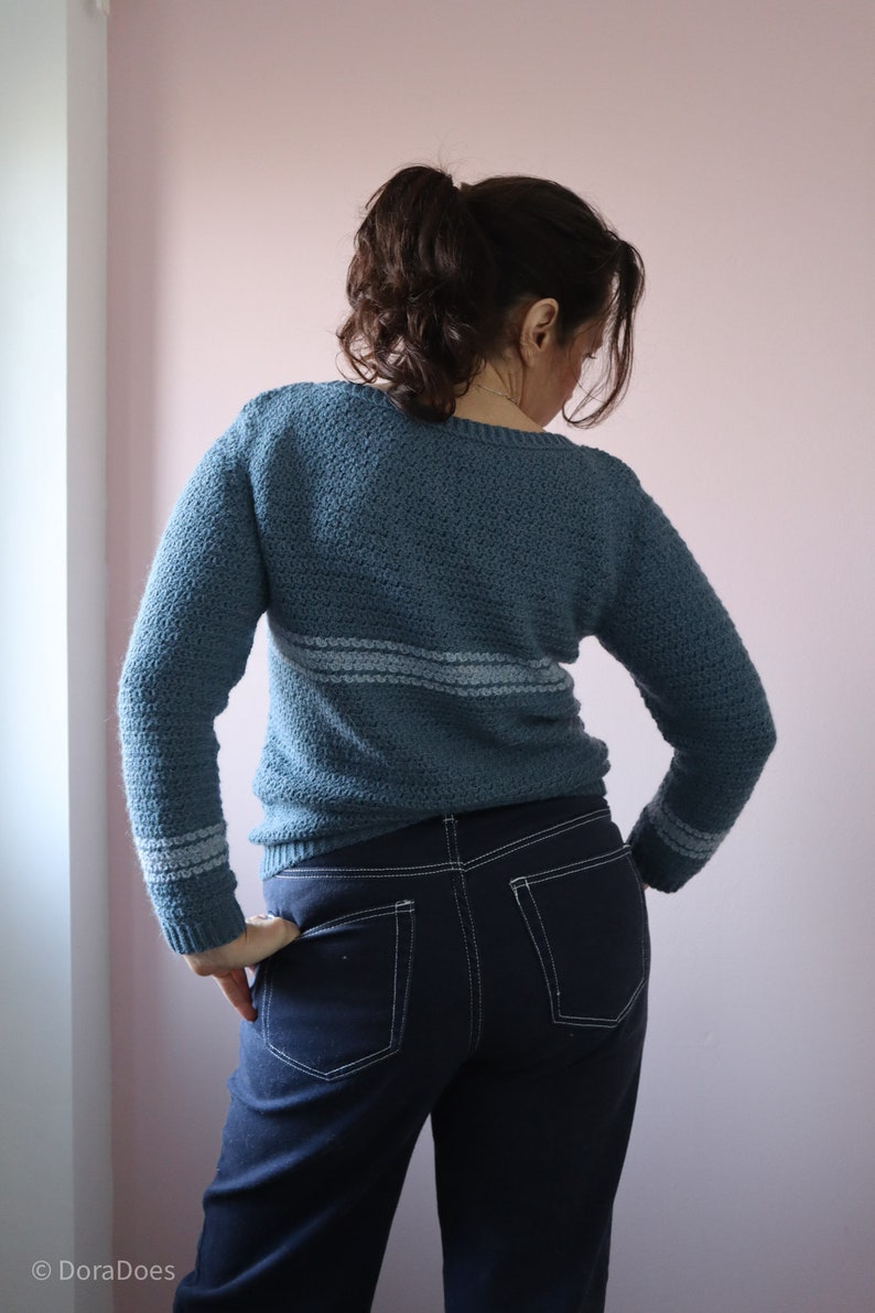 Dora is seen from behind wearing a blue long sleeve relaxed fit crochet sweater made from the Sequel sweater pattern. She has her hands on her hips, showing the stripes around the wrist.