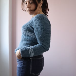 Dora seen from the side wearing a long sleeve relaxed fit modern crochet sweater paired with dark jeans.