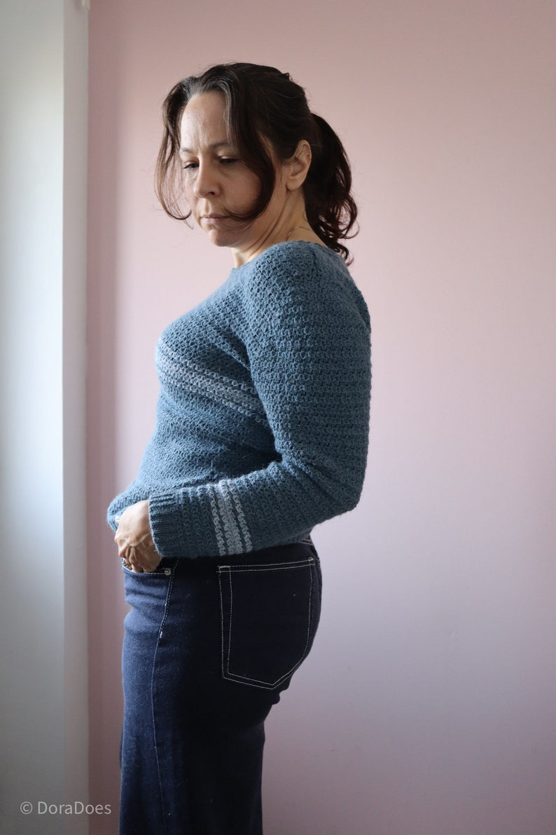 Dora seen from the side wearing a long sleeve relaxed fit modern crochet sweater paired with dark jeans.