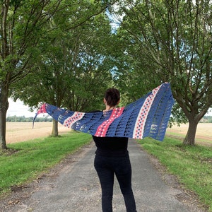 May include: A person wearing a blue, pink and white crocheted shawl with a geometric pattern. The shawl is draped over the person's shoulders and is flowing in the wind. The person is standing on a path in a field.