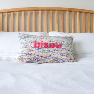 May include: A white bed with a wooden headboard and two white pillows. The top pillow has a colorful crocheted pillow with the word "bisou" in pink.