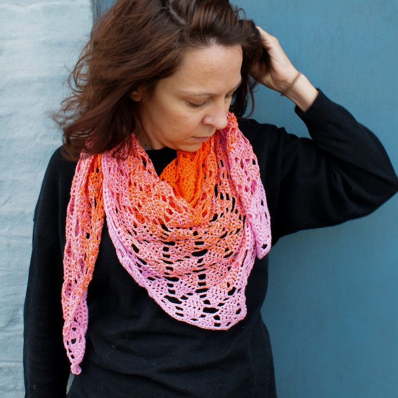 May include: A woman wearing a black sweater with a pink and orange crocheted shawl. The shawl has a delicate lace pattern and is draped around her neck.