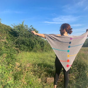 Peut inclure: Une personne portant un châle triangulaire gris avec des rayures et des cercles colorés. Le châle est fait d'un tissu doux et texturé. La personne se tient dans un champ avec de l'herbe verte et un ciel bleu en arrière-plan.